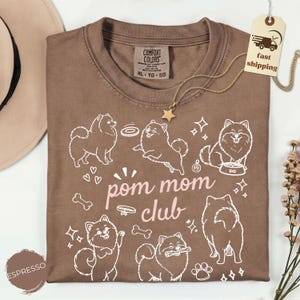 以下が含まれることがあります： 茶色のTシャツ。白いポメラニアン犬のグラフィックと「pom mom club」の文字がプリントされています。Tシャツはクルーネックで、「Comfort Colors」と書かれたタグが付いています。金の星のネックレスも見えます。
