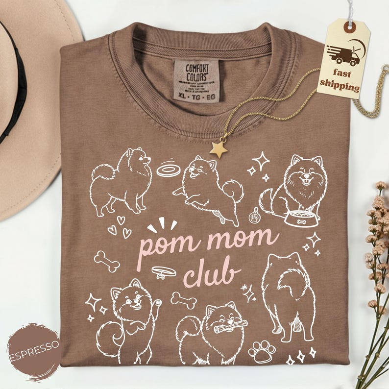 以下が含まれることがあります： 茶色のTシャツ。白いポメラニアン犬のグラフィックと「pom mom club」の文字がプリントされています。Tシャツはクルーネックで、「Comfort Colors」と書かれたタグが付いています。金の星のネックレスも見えます。