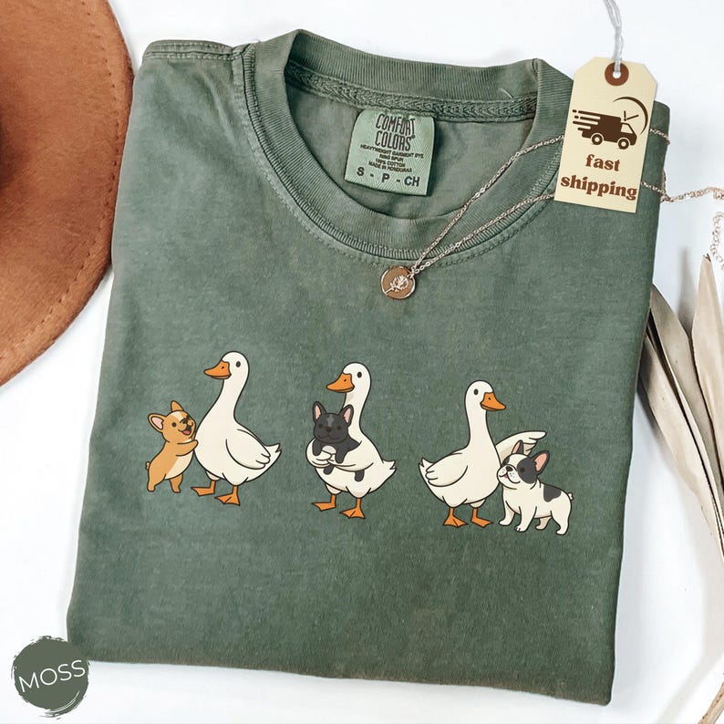 May include: Moss green Comfort Colors t-shirt featuring a graphic of three white geese with orange beaks and feet, each paired with a small dog. The dogs are a tan Chihuahua and two black and white French Bulldogs. A silver necklace is visible.