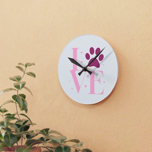 May include: A circular wall clock with a white face. The word "LOVE" is printed in pink, with a pink paw print above the "E". Black clock hands show the time. Small pink hearts are scattered around the letters. The clock is on a light orange wall.