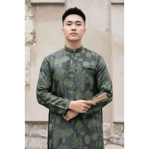 May include: A dark green tunic with a mandarin collar and long sleeves. The tunic features a repeating leaf pattern in a lighter green shade. The garment is worn by a person holding a small, light-colored fan.