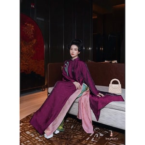 May include: A woman in a plum-coloured, floor-length dress with a high collar and long sleeves. The dress has a textured pattern and is paired with a pink and green underlay. A small, cream-coloured handbag sits on the sofa.