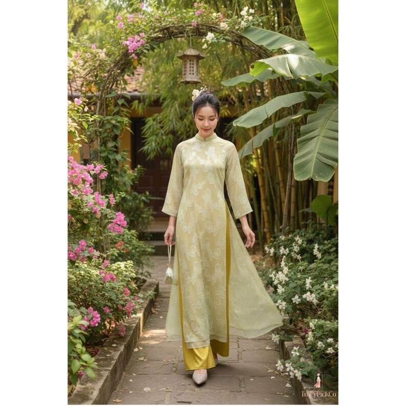 May include: A woman wearing a light yellow traditional dress with a high collar and long sleeves, walking on a stone path. The dress has a floral pattern and is paired with yellow pants. The background features lush greenery, flowers, and an archway.