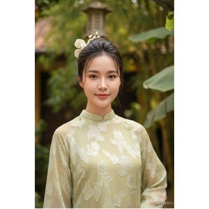 May include: A woman wearing a light green dress with white floral embroidery. The dress has long sleeves and a mandarin collar. She has a flower in her hair and is smiling. The background is blurred with greenery.