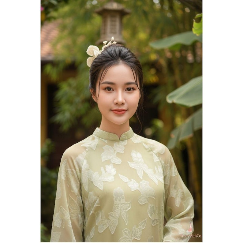 May include: A woman wearing a light green dress with white floral embroidery. The dress has long sleeves and a mandarin collar. She has a flower in her hair and is smiling. The background is blurred with greenery.