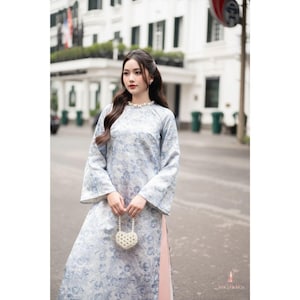 Könnte beinhalten: Ein hellblaues Kleid mit floralem Muster und langen, ausgestellten Ärmeln. Das Kleid hat einen hohen Ausschnitt und wird mit einer kleinen, herzförmigen, perlenbesetzten Handtasche kombiniert. Das Kleid wird im Freien auf einer Stadtstraße getragen.