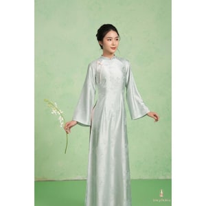 Könnte beinhalten: Ein hellgrünes, langärmeliges Kleid mit Stehkragen und floralem Muster. Das Kleid hat weite Ärmel und einen hohen Ausschnitt. Das Model hält einen weißen Blumenstiel. Der Hintergrund ist hellgrün.