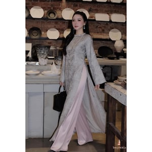 May include: A woman in a long, grey patterned tunic with wide sleeves and a high slit, paired with light pink palazzo trousers. She carries a black handbag. The background features a brick wall and shelves with decorative items.