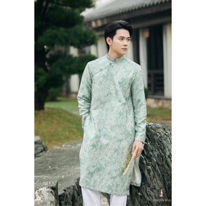 以下が含まれることがあります： 花柄とチャイナカラーの薄緑色のチュニック。長袖チュニックは白いパンツと組み合わせています。画像は、緑と建物のぼやけた背景のある屋外に設定されています。