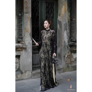 May include: A black lace dress with long sleeves and a high neckline. The dress features a floral pattern and a gold lining. A pearl necklace and a small black handbag complete the look. The dress is from TracyPickCo.