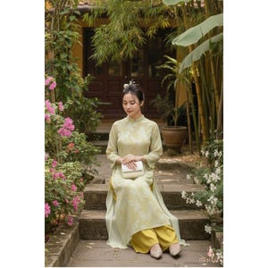May include: A woman wearing a light green traditional Vietnamese dress with a floral pattern, seated on stone steps. The dress has long sleeves and a high collar, paired with yellow pants. She holds a white clutch, with a background of lush greenery and flowers.