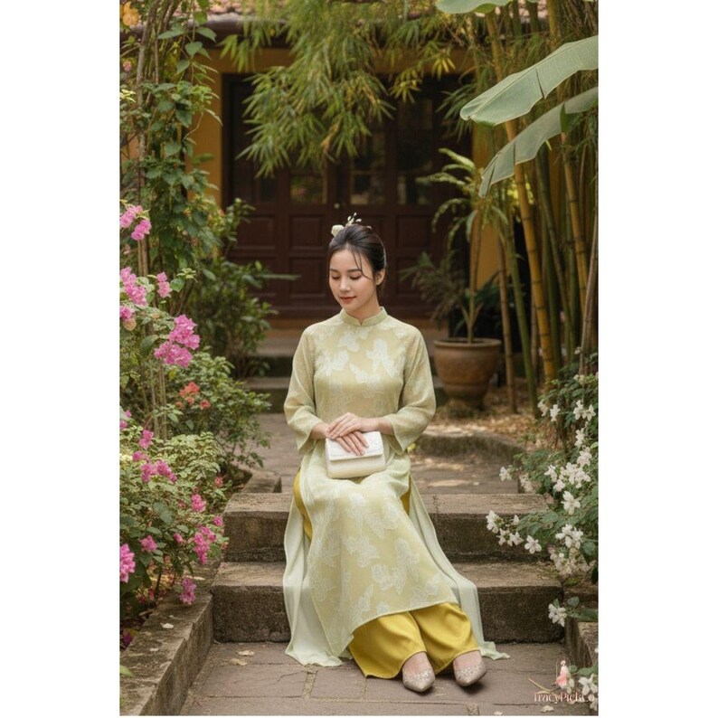 May include: A woman wearing a light green traditional Vietnamese dress with a floral pattern, seated on stone steps. The dress has long sleeves and a high collar, paired with yellow pants. She holds a white clutch, with a background of lush greenery and flowers.