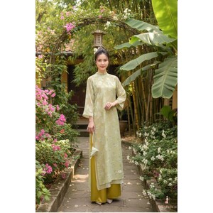 May include: A woman wearing a light green, long-sleeved dress with a floral pattern. The dress has a high collar and is paired with yellow pants. The setting is outdoors, with a floral archway and lush greenery in the background.