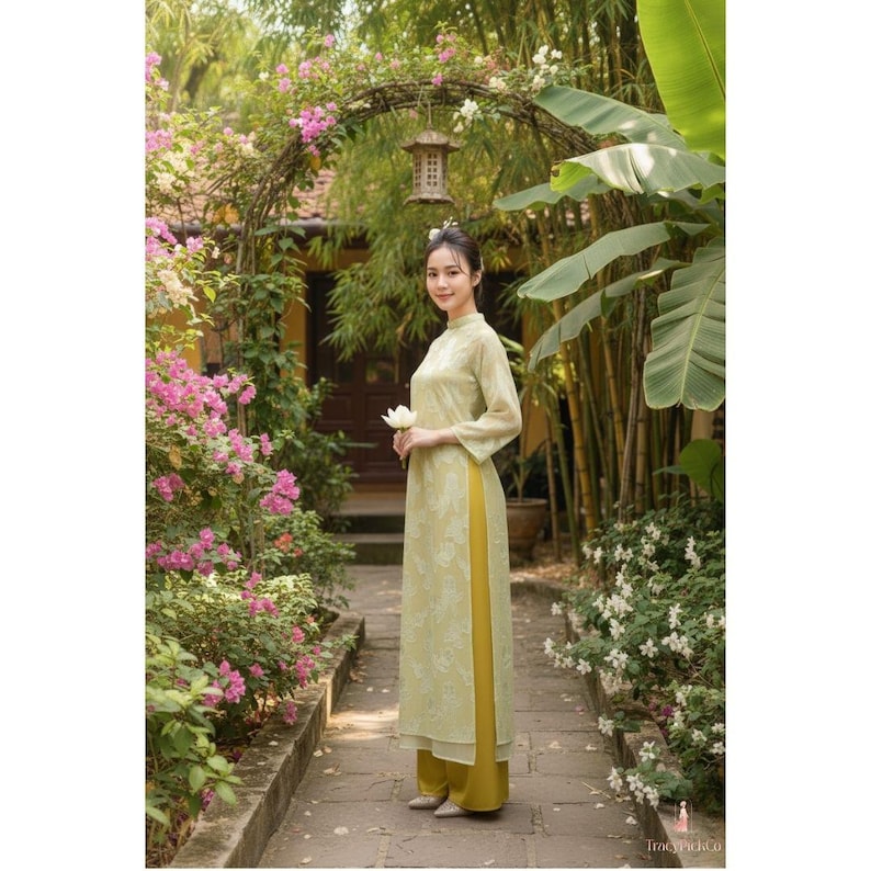 May include: A woman wearing a light green traditional Vietnamese dress, with a floral pattern, and yellow pants. She is holding a white flower and standing in a garden with pink flowers and bamboo.