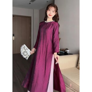 May include: A long, flowing magenta dress with long sleeves and a high slit. The dress has a subtle sheen and a white lace trim around the neckline. A white and silver fan is held in the hand.