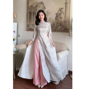 May include: A traditional Vietnamese Áo dài dress. The dress features a light gray floral pattern over a long tunic with a high collar and side slits. The dress is paired with wide-leg pink pants and white fabric.