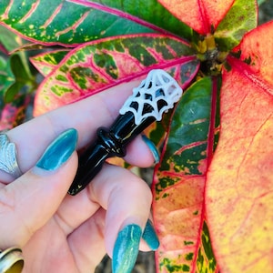 Puede incluir: Una pipa de vidrio negro con un diseño de telaraña blanca. La pipa se sostiene en una mano con esmalte de uñas azul verdoso.