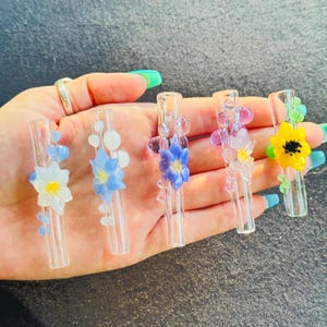 Puede incluir: Cinco tubos de vidrio transparente, cada uno con un diseño floral tridimensional único. Las flores incluyen pétalos blancos, azul claro, morados y amarillos con centros contrastantes, acentuados por pequeñas burbujas de vidrio.