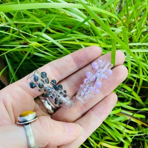 Könnte beinhalten: Zwei transparente Glaspfeifen, eine mit schwarzen und die andere mit lavendelfarbenen, blasenartigen Verzierungen, werden in einer Hand gehalten. Ein Silberring mit einem gelben Stein ist ebenfalls zu sehen. Der Hintergrund ist hellgrünes Gras.
