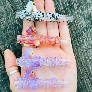 May include: Four glass pipes with textured, organic designs. The top pipe is white with black dots, the second is orange and red, the third is light purple, and the bottom is a darker purple. Each pipe features small raised glass accents and a clear mouthpiece.