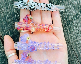 May include: Four glass pipes with textured, organic designs. The top pipe is white with black dots, the second is orange and red, the third is light purple, and the bottom is a darker purple. Each pipe features small raised glass accents and a clear mouthpiece.