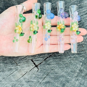 May include: Five clear glass smoking pipes, each adorned with colorful, decorative glass elements. The designs include green, yellow, and blue glass accents, with small, dark green turtle-like figures. The pipes are held in a hand against a weathered wood background.
