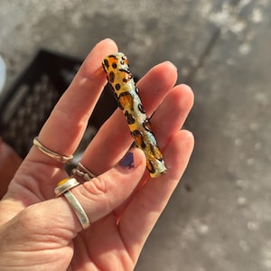 May include: A cylindrical, glass object with a leopard print design in shades of brown and amber. The object is held in a hand, and the background is a blurred, neutral color. The object appears to be a decorative item.