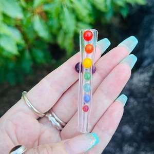 May include: A clear glass pipe featuring seven coloured spheres in a vertical arrangement. The spheres are red, orange, yellow, green, blue, and purple. The pipe is set against a backdrop of green foliage.