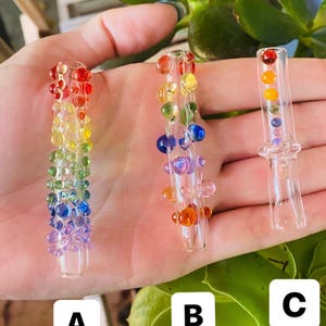 Puede incluir: Tres pipas de fumar de vidrio con detalles de cuentas de colores. La pipa A presenta un degradado arcoíris, la pipa B tiene cuentas azules, moradas y naranjas, y la pipa C tiene cuentas naranjas y amarillas. Las pipas se sostienen sobre un fondo de follaje verde.