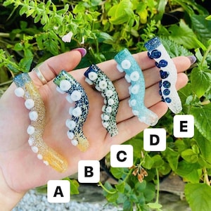 Pode incluir: Cinco tentáculos de polvo de vidro rotulados de A a E. O tentáculo A é dourado, B é azul escuro, C é verde, D é azul claro e E é branco com manchas azul escuro. Cada tentáculo apresenta ventosas brancas em relevo na superfície.