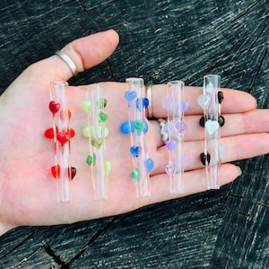 May include: Five clear glass straws, each decorated with small, raised heart-shaped glass accents in different colours. The set includes straws with red, light green, blue, purple, and black and white hearts arranged along the length of the glass.