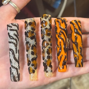 Può includere: Cinque unghie finte lunghe con vari disegni animalier. Le unghie presentano motivi come strisce zebrate, macchie leopardate e strisce tigrate nelle tonalità del nero, marrone, arancione e argento. Le unghie sono tenute in una mano.