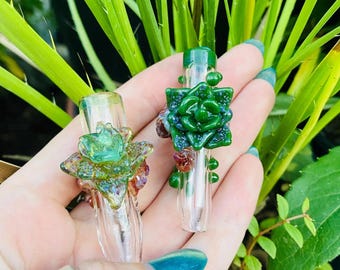 May include: Two clear glass pipes featuring decorative, sculpted glass succulent plant designs. One pipe displays a light green, textured succulent, while the other features a darker, vibrant green succulent with small, reddish-brown glass accents.