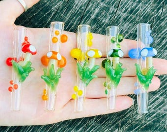 May include: Five clear glass tubes, each featuring a small sculpted mushroom with a colored cap and white spots, nestled in green glass foliage. The mushroom caps are red, orange, yellow, green, and blue, with matching colored glass dots on the tubes.