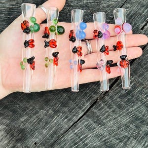 May include: A collection of six clear glass smoking pipes, each adorned with colorful glass beads and ladybug designs. The pipes feature green, blue, purple, and red accents, creating a whimsical and artistic aesthetic.