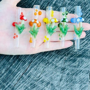 May include: Five clear glass straws, each adorned with a different colored mushroom and green leaf design. The mushrooms are red, orange, yellow, green, and blue. The straws are held in a hand.