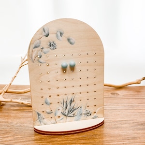 Könnte beinhalten: Ohrring-Präsentationsständer aus hellem Holz. Der Ständer hat Löcher für Ohrringe und ist mit bemalten botanischen Designs in Grau und Blau verziert. Zwei hellblaue Ohrringe sind ausgestellt. Der Ständer ist etwa 20 cm hoch.