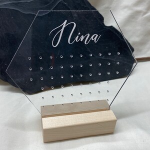 Könnte beinhalten: Ein transparenter sechseckiger Ohrringständer aus Acryl mit einem Holzständer. Der Ständer hat ein Gitter aus Löchern zum Aufbewahren von Ohrringen und der Name "Nina" ist oben eingraviert.