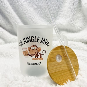 May include: A white frosted tumbler with a black and white logo that says "Lil Jungle Java" and a cartoon monkey holding a cup of coffee. The logo also says "Encinitas, CA". The tumbler has a bamboo lid with a hole for a straw. A clear plastic straw is included.