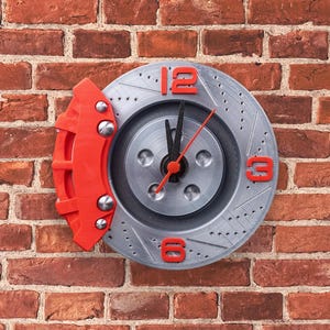 Peut inclure: Une horloge grise et rouge en forme de disque de frein de voiture. Le cadran de l'horloge a des aiguilles noires et des chiffres rouges. L'horloge est montée sur un mur en briques.
