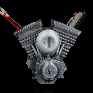 Puede incluir: Un organizador de escritorio de color plateado con forma de motor de motocicleta V2. El organizador contiene lápices y bolígrafos en los cilindros. La base del motor tiene el texto "V2" en relieve. El fondo es negro.