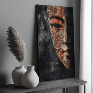 Puede incluir: Una obra de arte enmarcada que presenta un rostro parcial con intrincados jeroglíficos naranjas sobre un fondo oscuro. La obra está sobre una mesa gris, junto a dos jarrones blancos con hierba de la pampa decorativa.