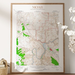 May include: Framed vintage map print of Moab, Utah, with text "MOAB" at the top. The map features a detailed topographical layout with green, brown, and red colorations. The print is in a wooden frame, displayed on a wooden surface.