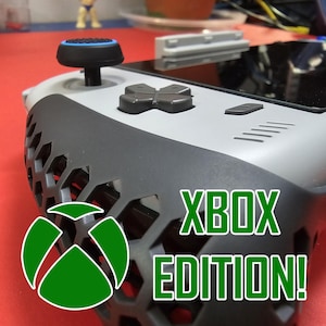 Może przedstawiać: Szary i czarny przenośny sprzęt do gier z czarnym joystickiem i zielonym logo Xbox. Urządzenie ma czarny i szary uchwyt w kształcie plastra miodu i napis "XBOX EDITION!" nadrukowany na boku.