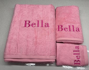 Puede incluir: Un juego de tres toallas de baño rosas, cada una bordada con el nombre "Bella" en hilo rosa más oscuro. El juego incluye una toalla de baño grande, una toalla de mano y un paño.