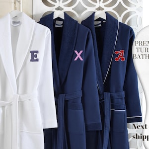 May include: Three premium Turkish bathrobes hanging on white hangers. One white robe with a dark blue embroidered "E" and two navy blue robes with embroidered letters. The text "Premium Turkish Bathrobe" is visible.