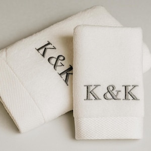 May include: Two white cotton towels featuring a textured border and the embroidered letters K&K in a dark grey serif font. The larger towel is folded horizontally, while the smaller towel is folded vertically, both displaying the monogram.