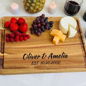 May include: A wooden serving board with a personalized inscription "Oliver & Amelia EST 10.20.2000". The board is filled with various snacks, including grapes, strawberries, cheese, and pineapple.