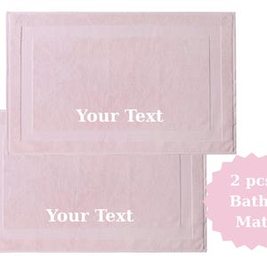 May include: Two light pink bath mats with a rectangular design and a white border. The text "Your Text" is printed on each mat. A pink starburst graphic reads "2 pcs Bath Mat".