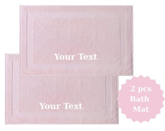 May include: Two light pink bath mats with a rectangular design and a white border. The text "Your Text" is printed on each mat. A pink starburst graphic reads "2 pcs Bath Mat".